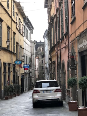 Une rue étroite avec une voiture garée, des bâtiments à l’architecture traditionnelle et une enseigne indiquant « Olivetti Associazione Pagani ».