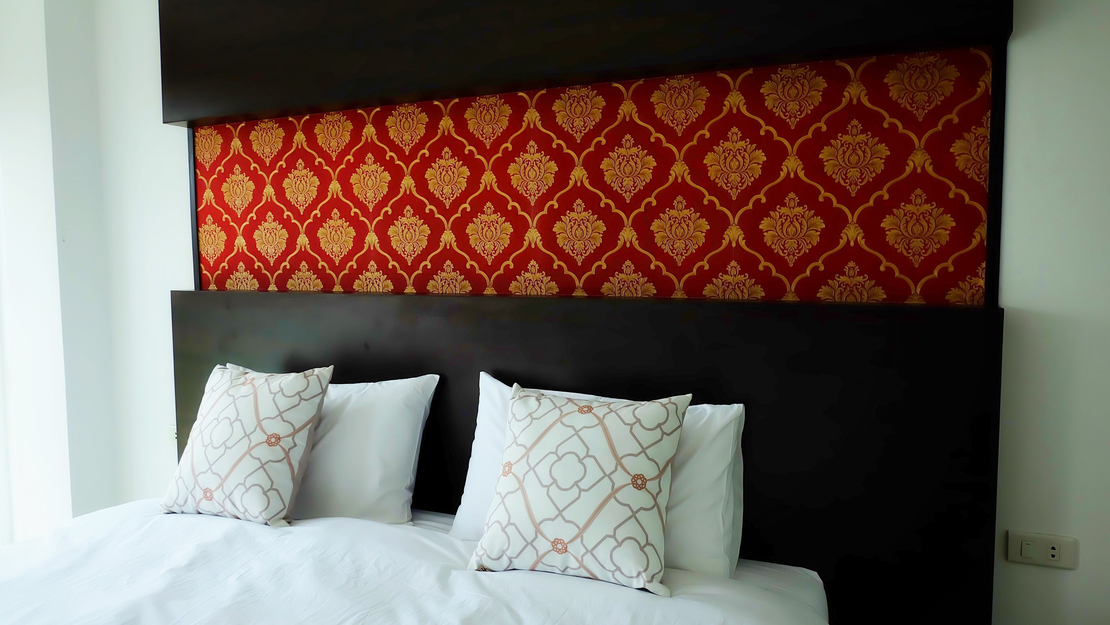 A bed with white linens and patterned pillows, a dark headboard, and a red patterned wall above.