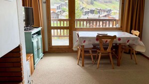 A room with a wooden table and chairs, a television on a cabinet, and a large window with a view of a village and mountains. - Les Côtes 15 - 2-room apartment with mountain view (LES AVANCHERS VALMOREL)