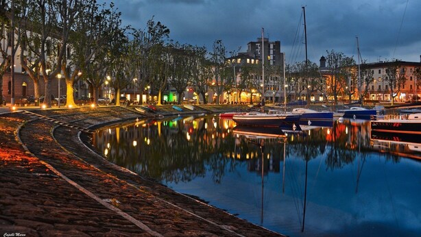 Une zone riveraine où sont amarrés des bateaux, avec un canal et des bâtiments dont les fenêtres sont illuminées.