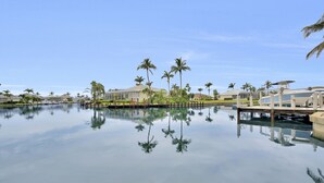 A calm water body reflecting palm trees and buildings, with a clear sky above. - WIDE-VIEW ESTATE HOME NEAR BOAT RAMP WITH POOL & DEEP-WATER DOCKAGE (Marco Island)