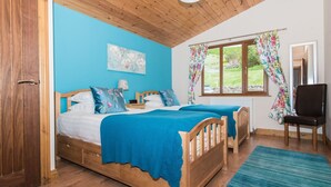 Ein Schlafzimmer mit zwei Holzbett, einer blauen Wand, einem Fenster mit floralen Vorhängen und einem Holzstuhl.