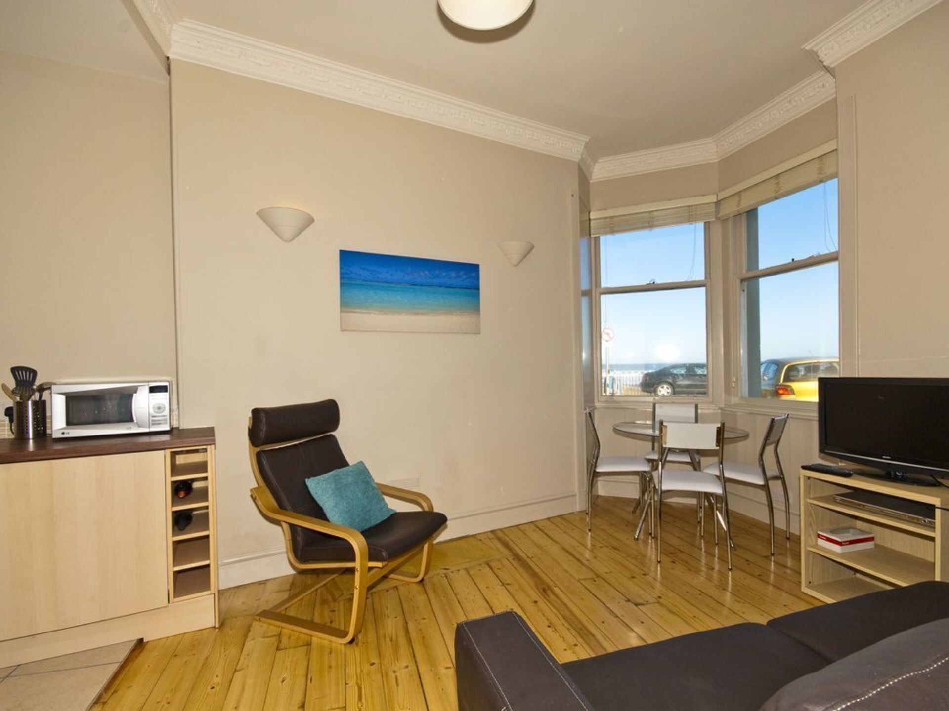 A compact living space with a wooden floor, a kitchenette with a microwave, a black leather chair with a blue pillow, a TV on a wooden stand, and a painting of a beach on the wall.
