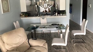 Un coin repas avec une table en verre, des chaises blanches, un lustre et une cuisine visible à travers une porte ouverte.