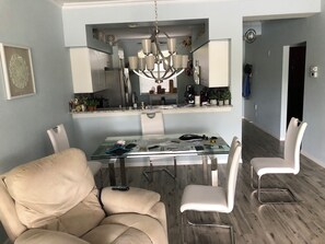 Zona pranzo con tavolo in vetro, sedie bianche, lampadario e cucina visibile attraverso un'apertura.
