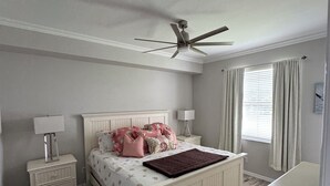 Une chambre à coucher avec un lit, des tables de chevet, un ventilateur de plafond et une fenêtre avec des rideaux.