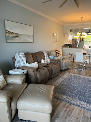 A living room with a sectional sofa, a coffee table, and a rug. There is a kitchen area with a dining table and chairs.