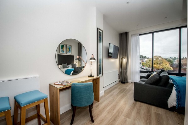 A modern living room with a wooden desk, a round mirror, a blue chair, a grey sofa, and a large window offering a city view. - The James Suites (Londonderry)