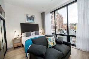 A modern hotel room with a large bed, a sofa, a nightstand, and a view of the city through large windows. - The James Suites (Londonderry)
