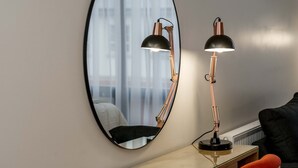 A room with a round mirror reflecting a bed and a window with curtains. There is a desk with a lamp and a chair. - The James Suites (Londonderry)