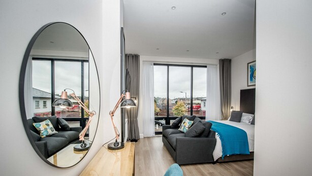A modern hotel room with a large mirror, two sofas, a bed, and a view of the city. - The James Suites (Londonderry)