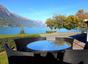 A round table with wicker chairs, a bench, and a view of a lake with mountains in the background. - Apartment 1.  Lakeside property.   Amazing Views, balcony & garden. (Bönigen b. Interlaken)