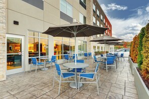 An outdoor patio with tables and chairs, a building with large windows, and a car parked nearby.