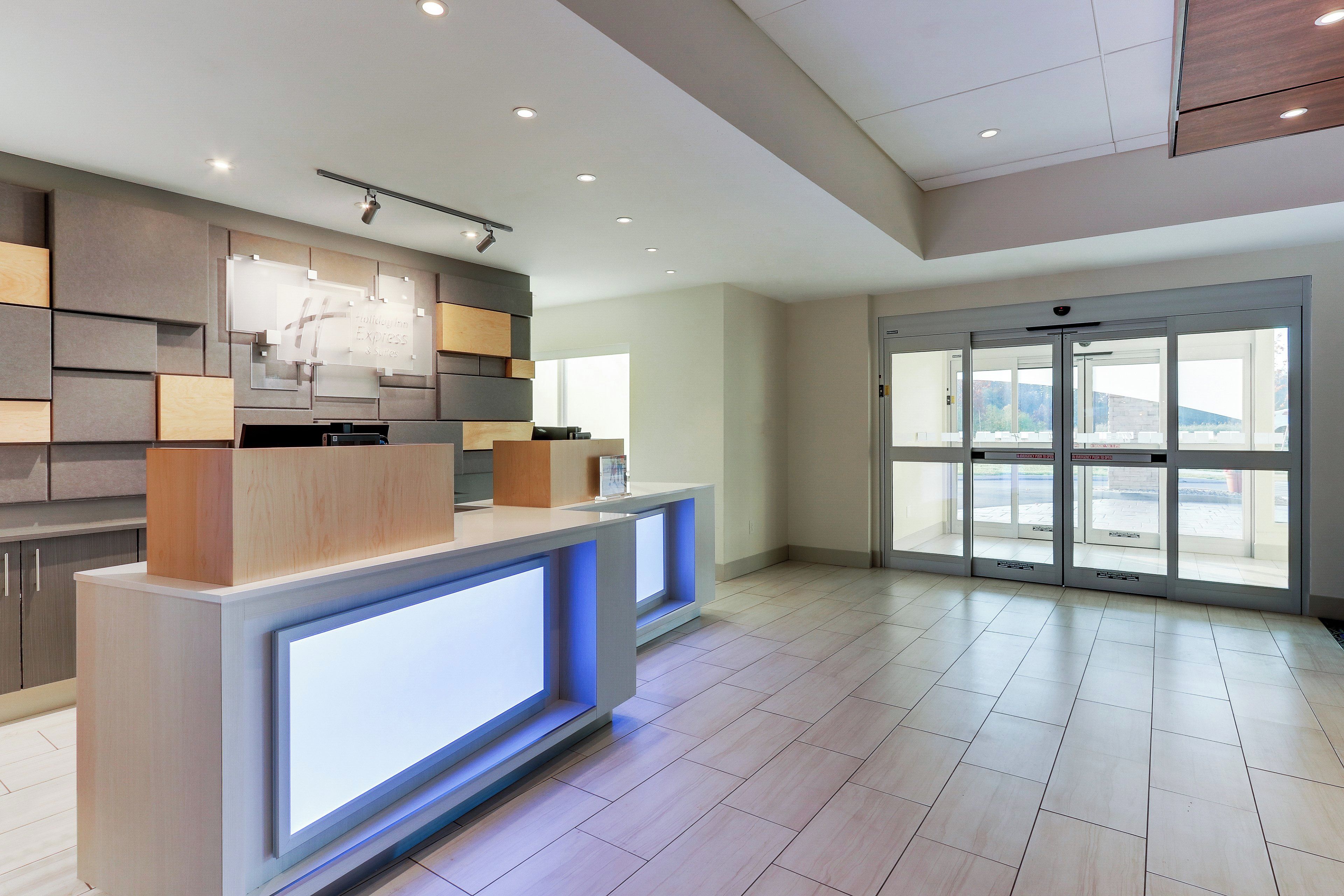 A hotel reception area with a modern design, featuring a reception desk, a digital display, and a view through the glass doors.