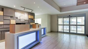 A hotel reception area with a modern design, featuring a reception desk, a digital display, and a view through the glass doors.