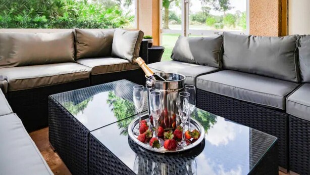 A modern outdoor seating area with a glass-top table, a champagne bucket, and glasses, surrounded by cushioned sofas.