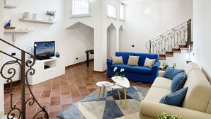 A living room with a blue sofa, a white coffee table, and a TV on a white stand. There is a staircase with a wrought iron railing leading to an upper floor. - Villa Lia (Sant'Agnello)