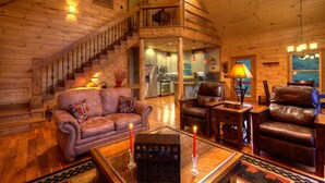 A cozy log cabin interior with a wooden staircase, a living room with a sofa and armchairs, a dining area, and a kitchen.