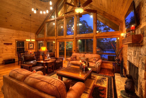 A cozy log cabin interior with a fireplace, leather furniture, and large windows offering a view of the outdoors.
