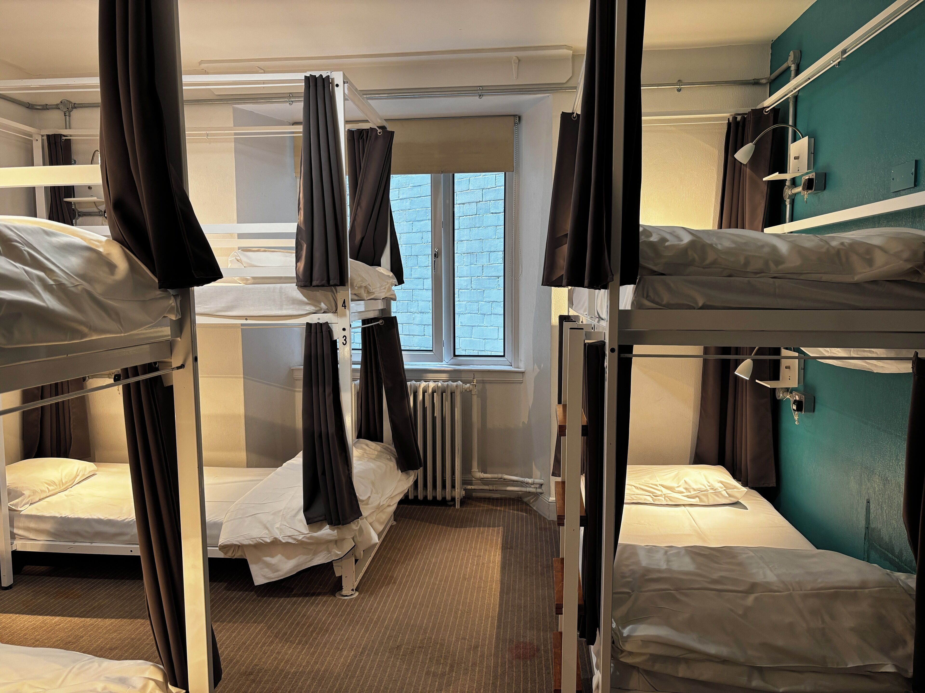 A dormitory room with bunk beds, a window, a radiator, and a desk with a lamp.