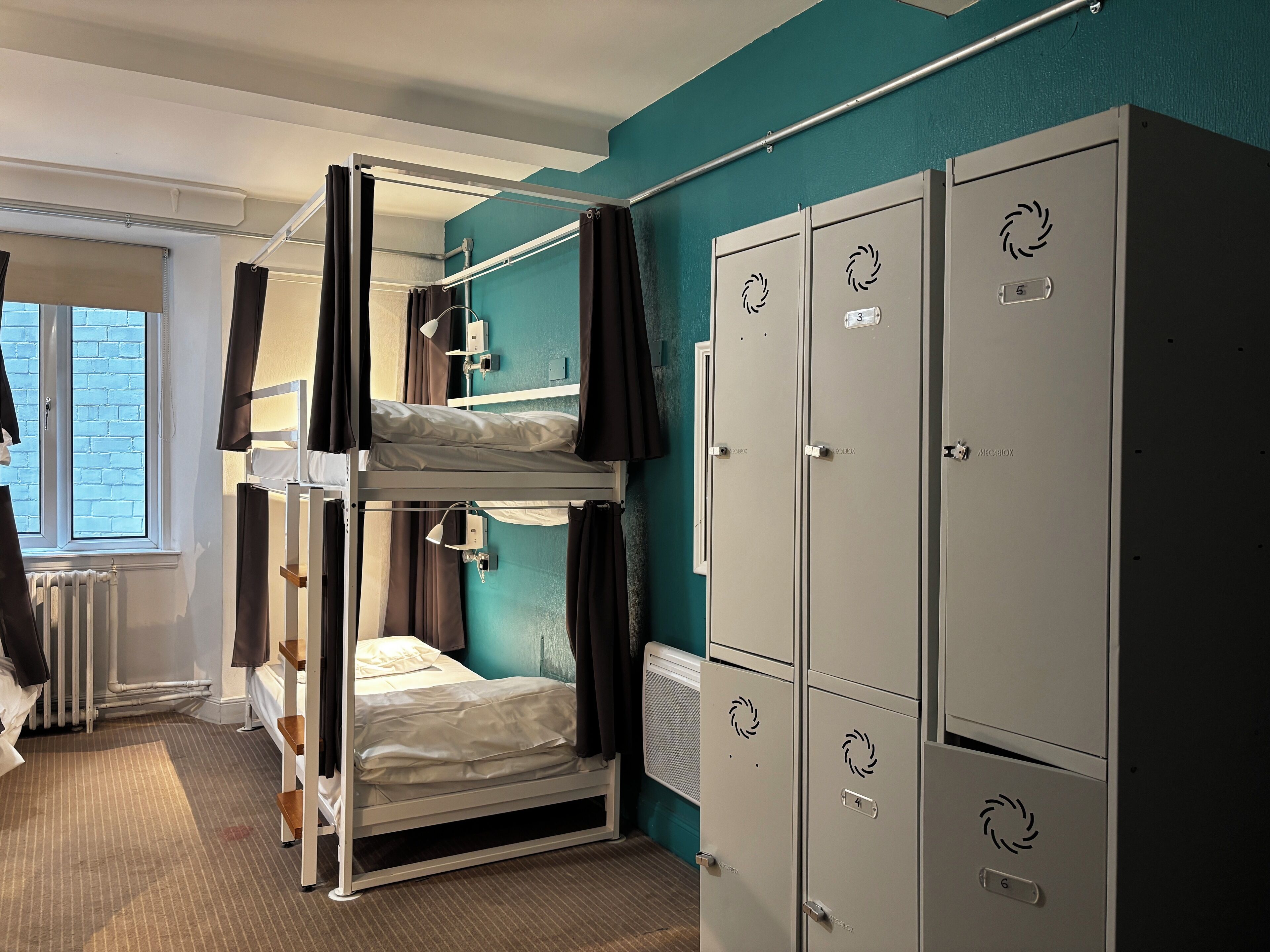 A dormitory room with bunk beds, a window with blinds, a radiator, and a set of lockers.