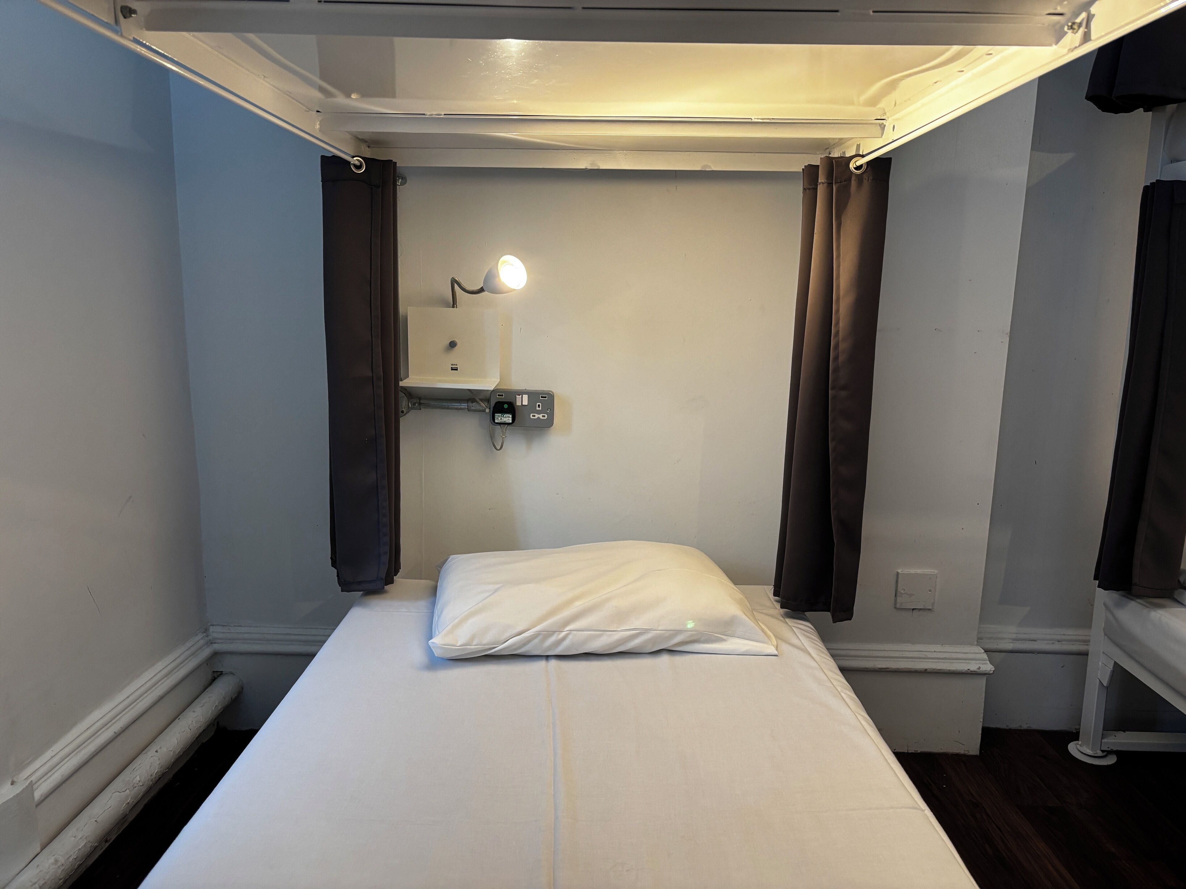A small, narrow room with a single bed, a bedside table, a wall-mounted light, and a small shelf with a power outlet.