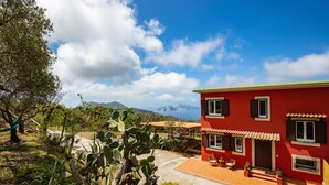 A red building with a tiled roof, a patio with outdoor furniture, and a view of mountains in the background. - Casa Aprea (Massa Lubrense)