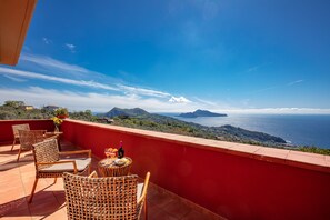Una terraza con sillas de mimbre y una mesa, con vistas a un paisaje costero con montañas y el mar.