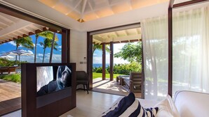 A modern hotel room with a large TV, a bed with striped pillows, a balcony with a view of palm trees and the ocean, and a patio area with outdoor seating. - Villa MAISON BLANC BLEU – Iconic Elegance Above Gouverneur (Gustavia)
