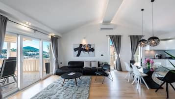 A modern living room with a glass dining table, black and white chairs, a black sofa, and a large window with a view of a cityscape.