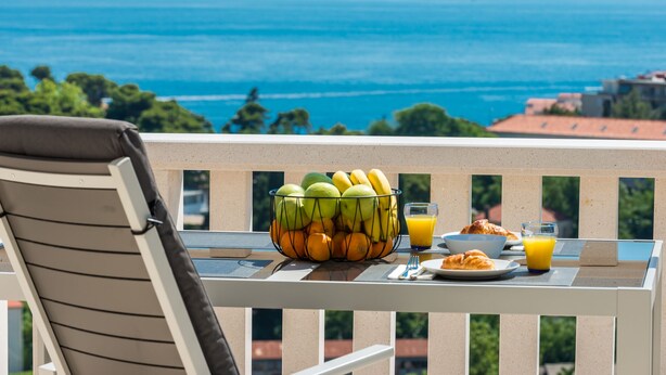 A balcony with a table set for breakfast, including fruits, croissants, and orange juice, overlooking a coastal view.