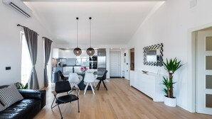 A modern living room with a black leather sofa, a dining area with a white table and chairs, a kitchen with stainless steel appliances, and a large mirror on the wall.