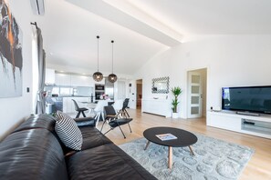 Uma sala de estar moderna com sofá de couro, mesa de centro e televisão. Há um espaço de refeições com cadeiras e uma cozinha ao fundo.