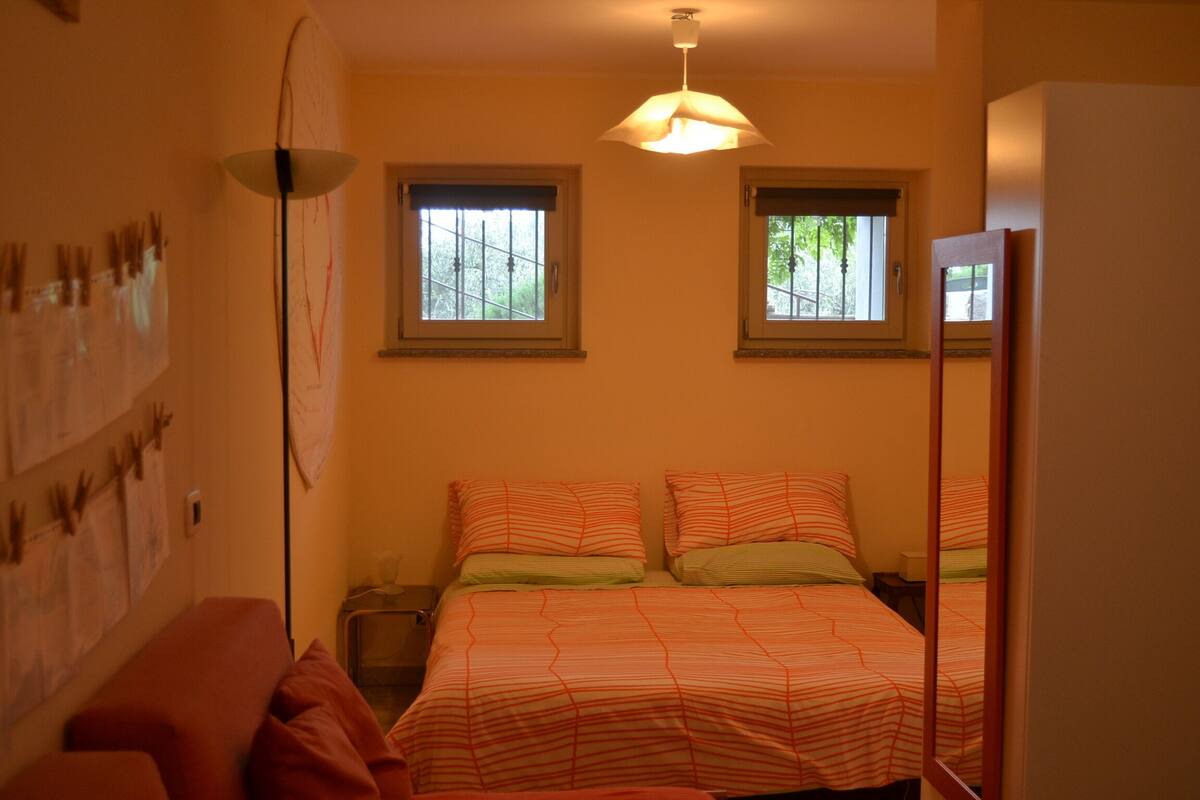 Un dormitorio con cama, dos ventanas, una lámpara de pie y un estante en la pared.