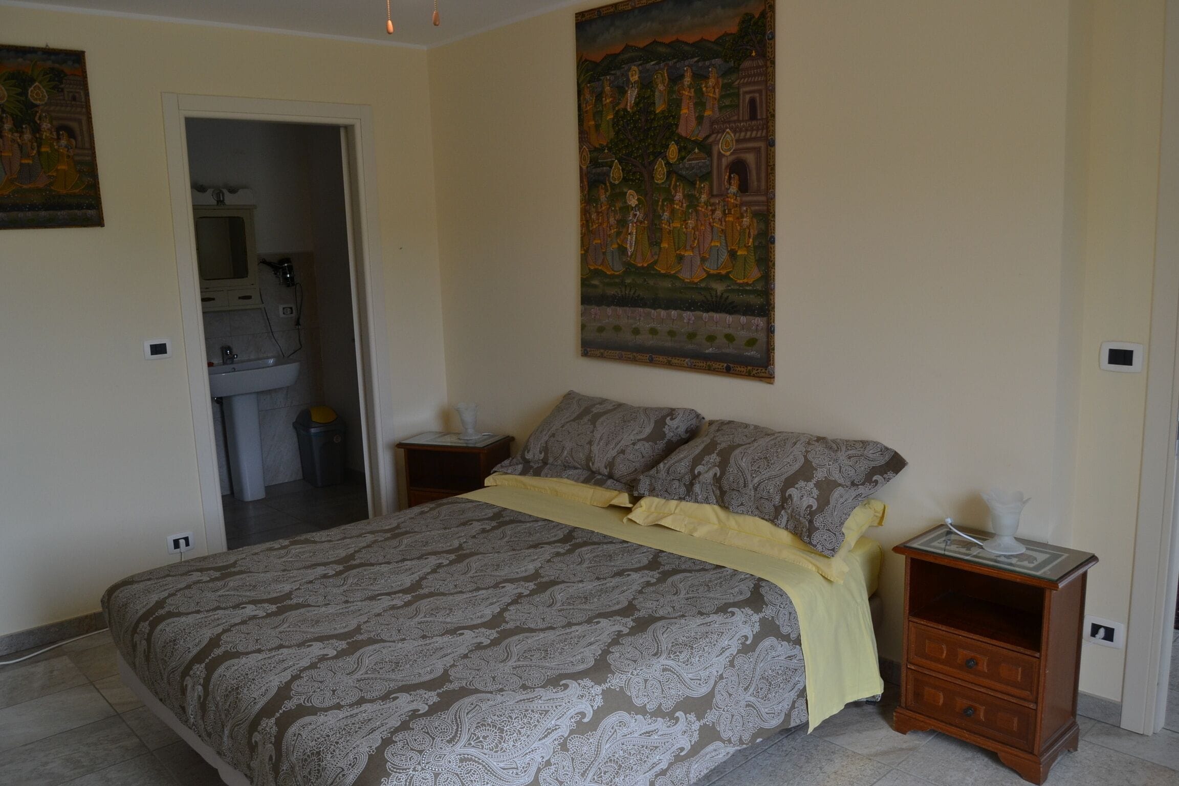 Une chambre à coucher avec un lit, une table de chevet, un tableau et une salle de bain visible à travers une porte ouverte.