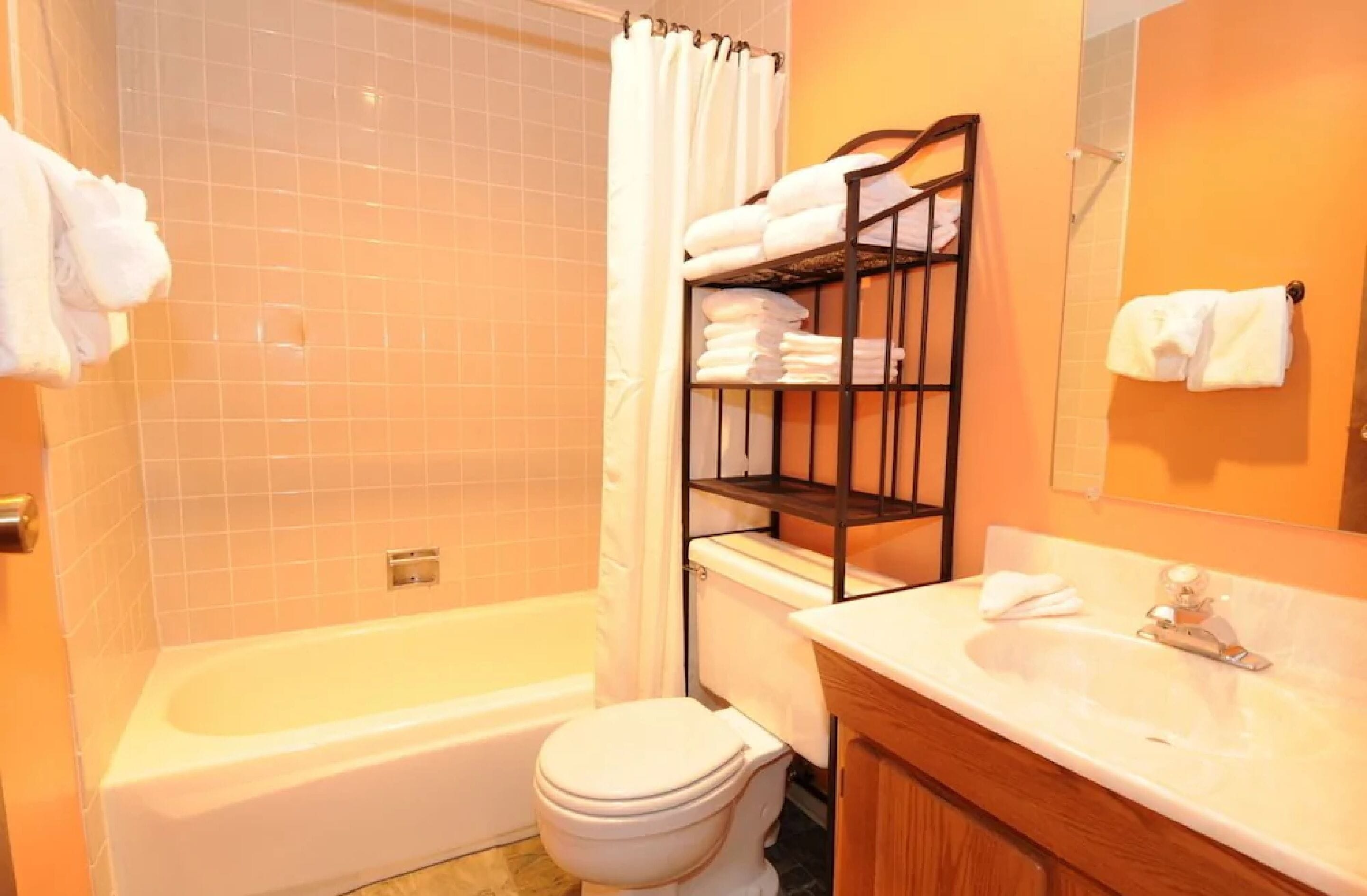 A bathroom with a bathtub, toilet, and sink. There is a towel rack with folded towels and a shelf with more towels.