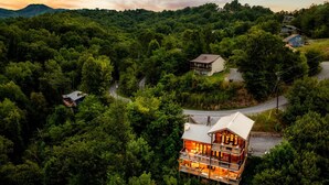 A multi-story wooden cabin with a balcony, surrounded by dense forest and a winding road.