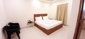 A hotel room with a single bed, bedside table, and a nightstand. There are two chairs and a window with curtains.