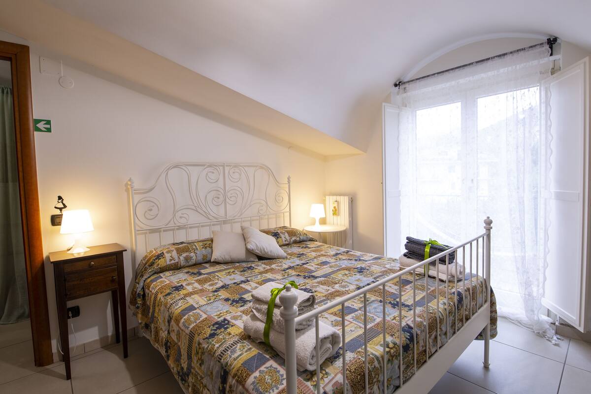 A neatly made bed with a patterned bedspread, two bedside tables with lamps, and a window with sheer curtains.