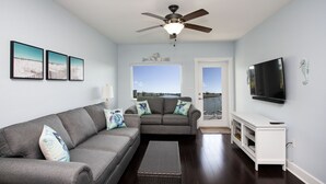 A living room with a grey sofa, a white console table, a flat-screen TV, and a ceiling fan. - Waterfront Condo+4 FREE Waterpark Passes-Walk to Beach-Family Favorite-Sleeps 8 (Indian Rocks Beach)
