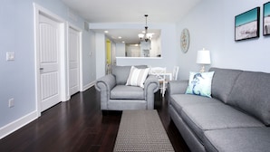 A living room with a grey sofa, a grey armchair, and a patterned rug. There is a white door on the left and a white wall with two framed pictures. - Waterfront Condo+4 FREE Waterpark Passes-Walk to Beach-Family Favorite-Sleeps 8 (Indian Rocks Beach)