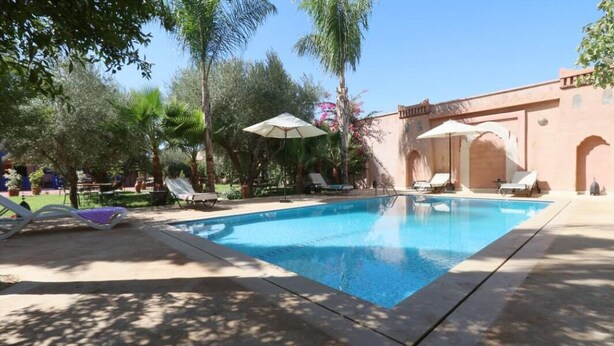 A swimming pool with lounge chairs and umbrellas, surrounded by palm trees and a building with arches. - Le Bleu House (Sidi Abdallah Ghiat)