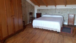 Un dormitorio con techo de madera, cama con colcha blanca, armario y pared de piedra.