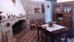 Un comedor con piso a cuadros, chimenea de piedra, sillas de madera, una mesa con mantel de encaje y un aparador con puertas de vidrio.