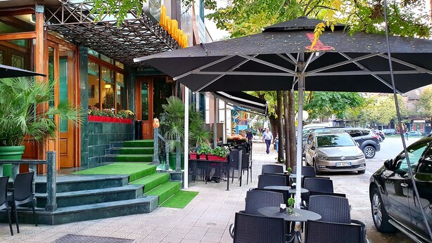 An outdoor cafe with outdoor seating, a staircase leading to the entrance, and a car parked nearby. - Nov Hotel (Tirana)