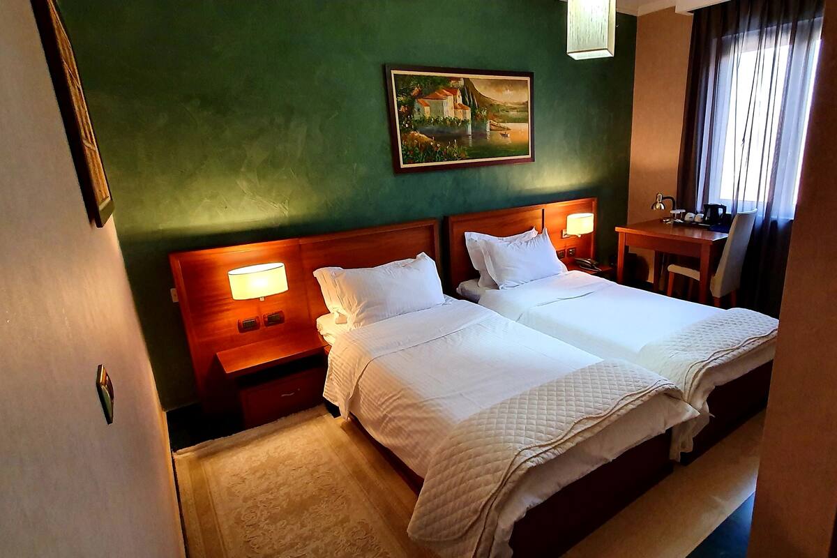 A hotel room with two beds, a desk, a chair, a painting on the wall, and a window with curtains.