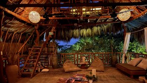 A cozy, rustic room with a thatched roof, wooden furniture, and a view of lush greenery.
