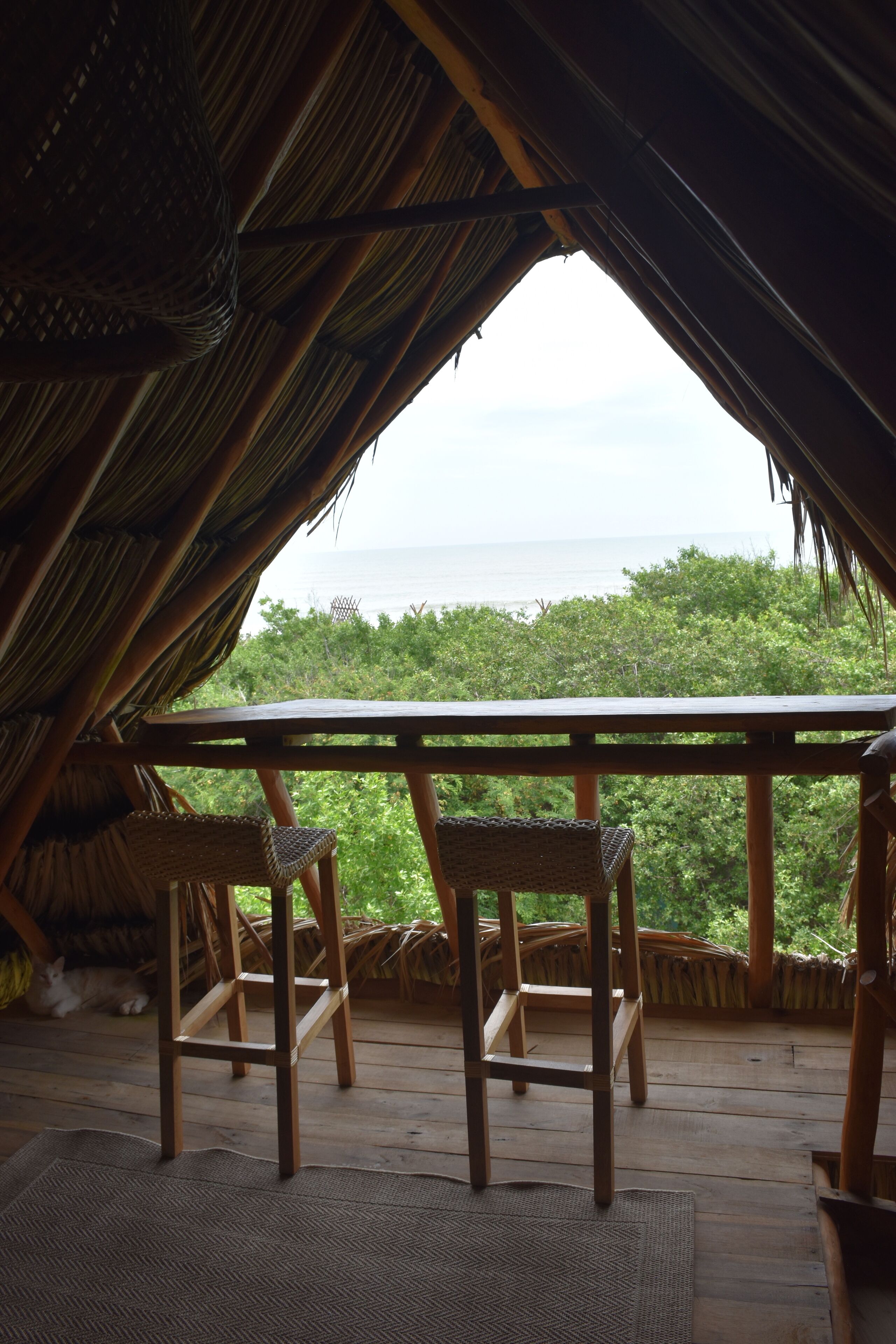 Een houten terras met twee rieten stoelen en een tafel, met uitzicht op een weelderig groen landschap en de zee.