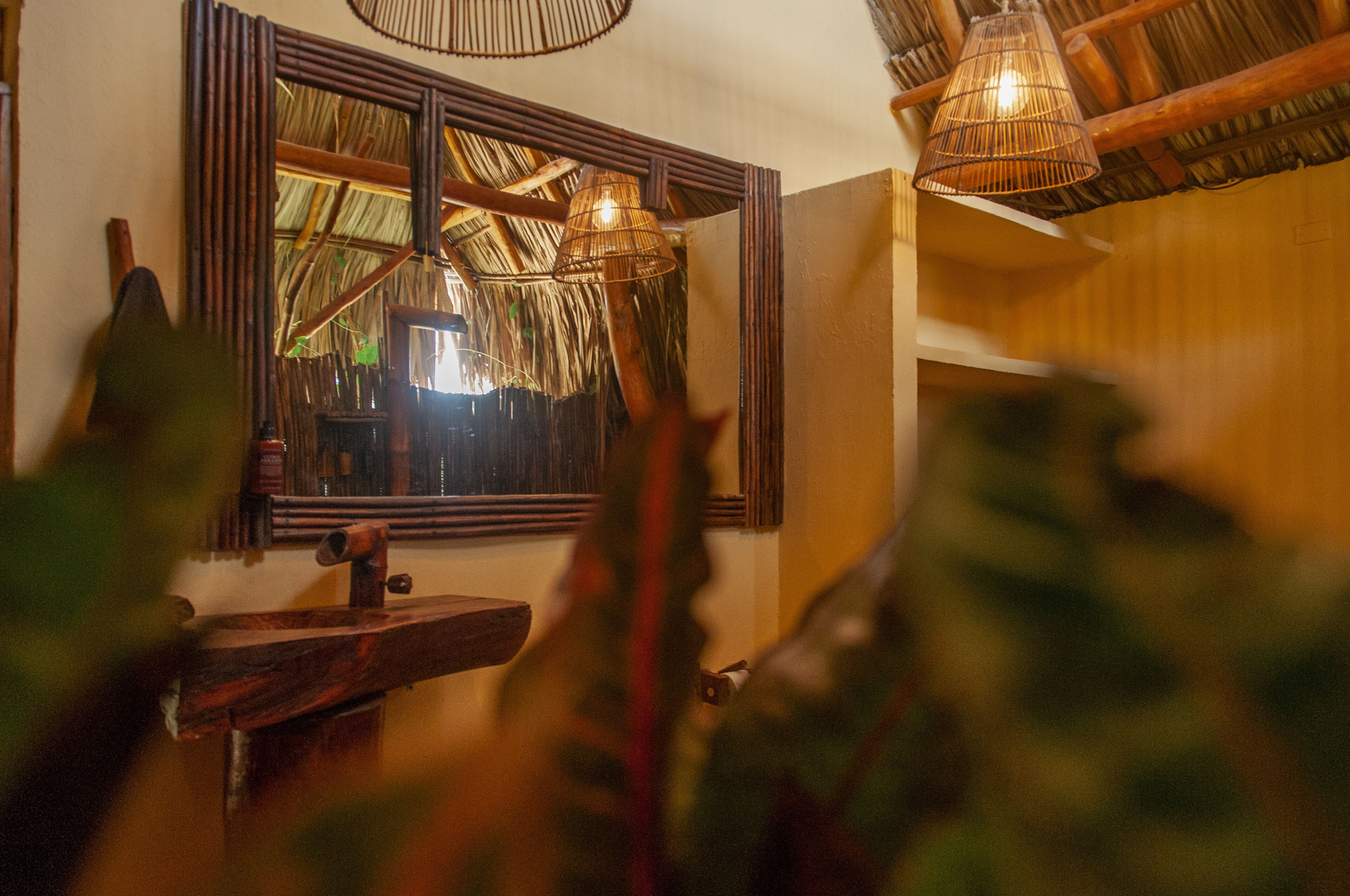 A rustic bathroom with a wooden sink, a large mirror, and a thatched roof visible through the window.