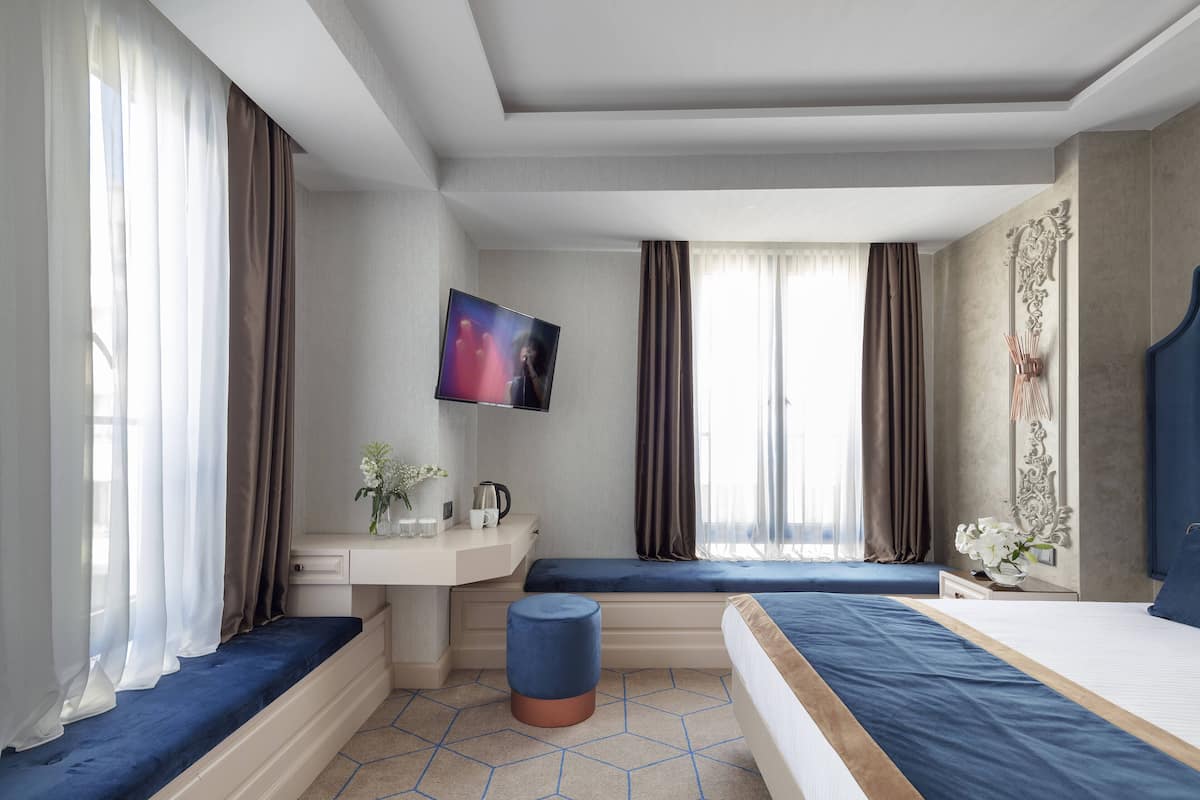 Una habitación de hotel moderna con una cama grande, un escritorio con silla, un taburete pequeño y un televisor montado en la pared.